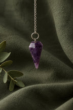 A faceted purple crystal cone pendant necklace hanging from a silver chain, displayed on a textured light gray fabric surface with a floral candle and green leaves in the background.