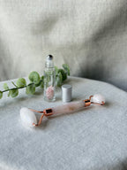 Rose quartz facial roller tool with rose gold metal accents, displayed on a light gray fabric surface next to a clear glass roller bottle with pink salt and a silver cap, with green eucalyptus leaves in the background.