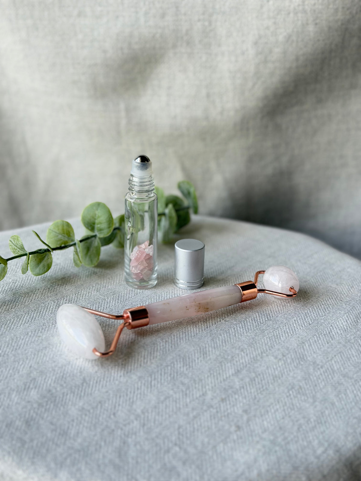 Rose quartz facial roller tool with rose gold metal accents, displayed on a light gray fabric surface next to a clear glass roller bottle with pink salt and a silver cap, with green eucalyptus leaves in the background.