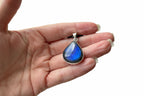 A hand holding a teardrop-shaped labradorite pendant with a silver bezel setting, showcasing its deep blue color and natural play-of-color, with a home interior background.