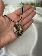 A hand holding a black cord necklace with an oval brown and beige spotted stone poppy jasper pendant against a white satin fabric background.