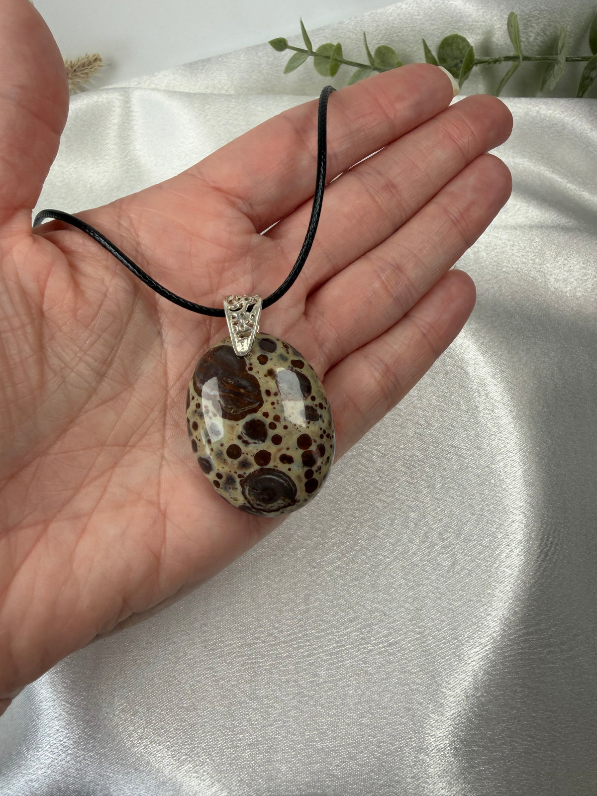 A hand holding a black cord necklace with an oval brown and beige spotted stone poppy jasper pendant against a white satin fabric background.