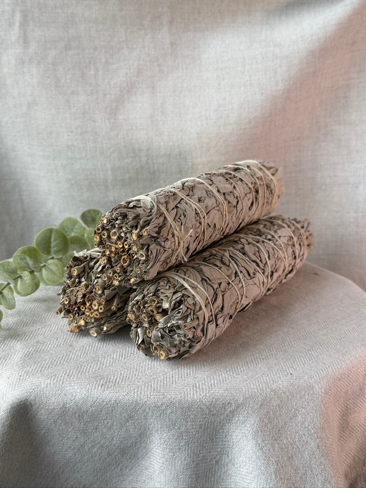 Two dried sage smudge sticks with green eucalyptus leaves, resting on a draped light beige linen fabric background.
