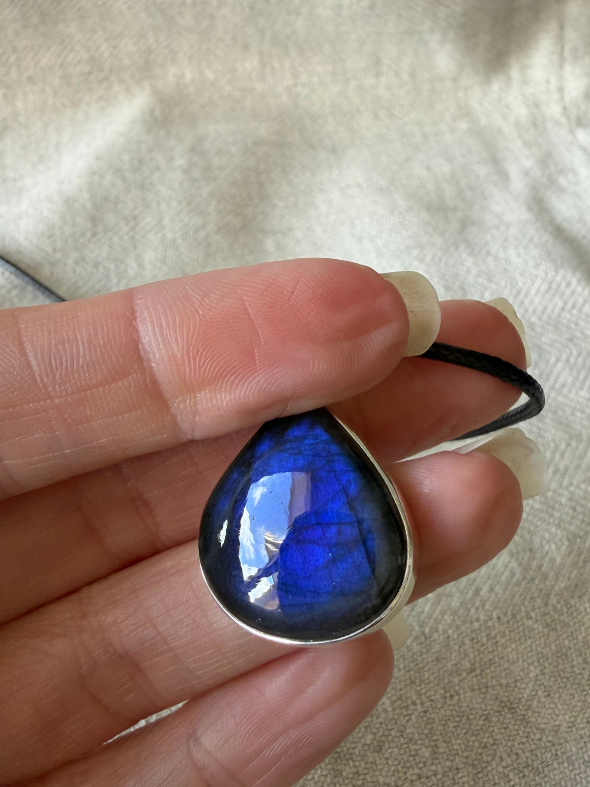 A hand holding a teardrop-shaped blue labradorite pendant in a silver bezel setting, attached to a black cord, against a beige linen fabric background.