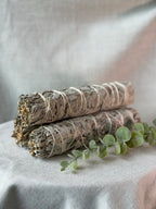Two bundled white sage smudge sticks wrapped in natural twine, arranged on a light fabric surface with a sprig of green eucalyptus in the foreground.