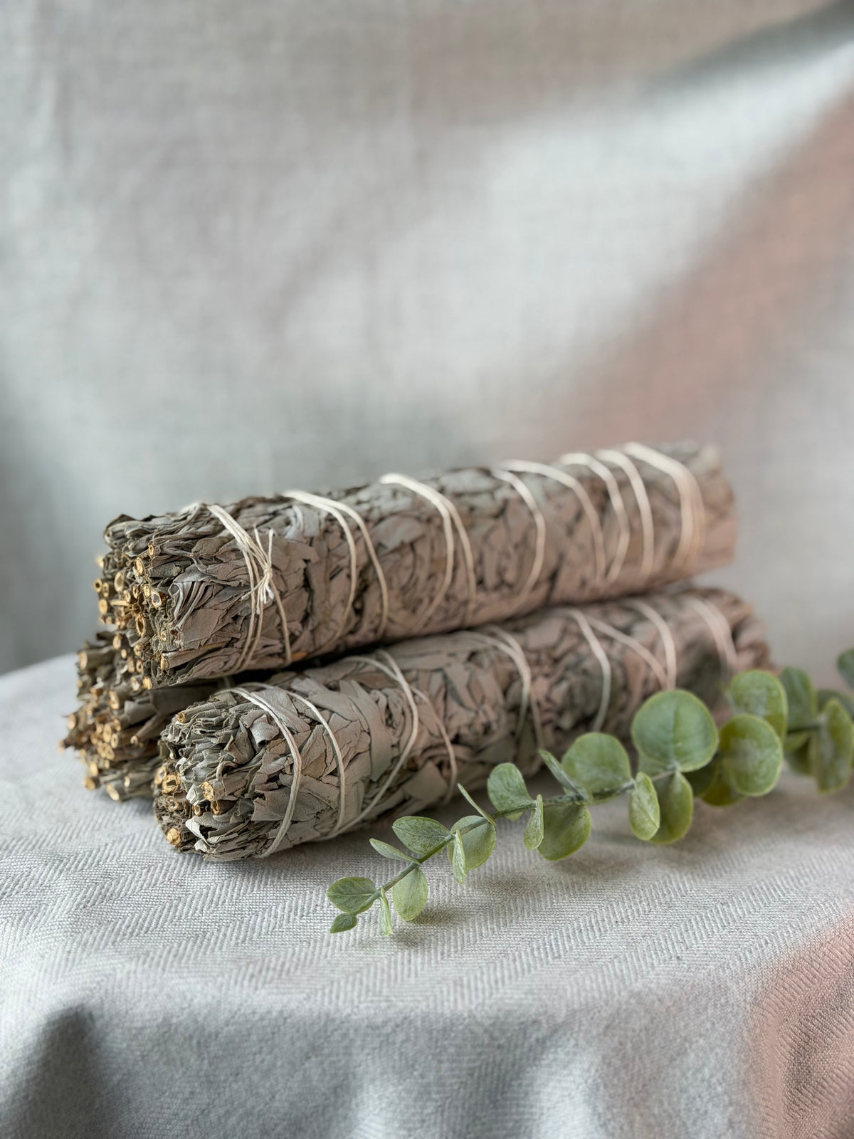 Two bundled white sage smudge sticks wrapped in natural twine, arranged on a light fabric surface with a sprig of green eucalyptus in the foreground.