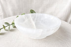 A translucent white crystal bowl made of what appears to be selenite or quartz, placed on a textured beige fabric surface with a green sprig of leaves as a prop.