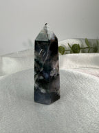 A natural pointed crystal tower, likely amethyst, standing upright on a draped white fabric surface with a soft gray background and a small green plant in the distance.