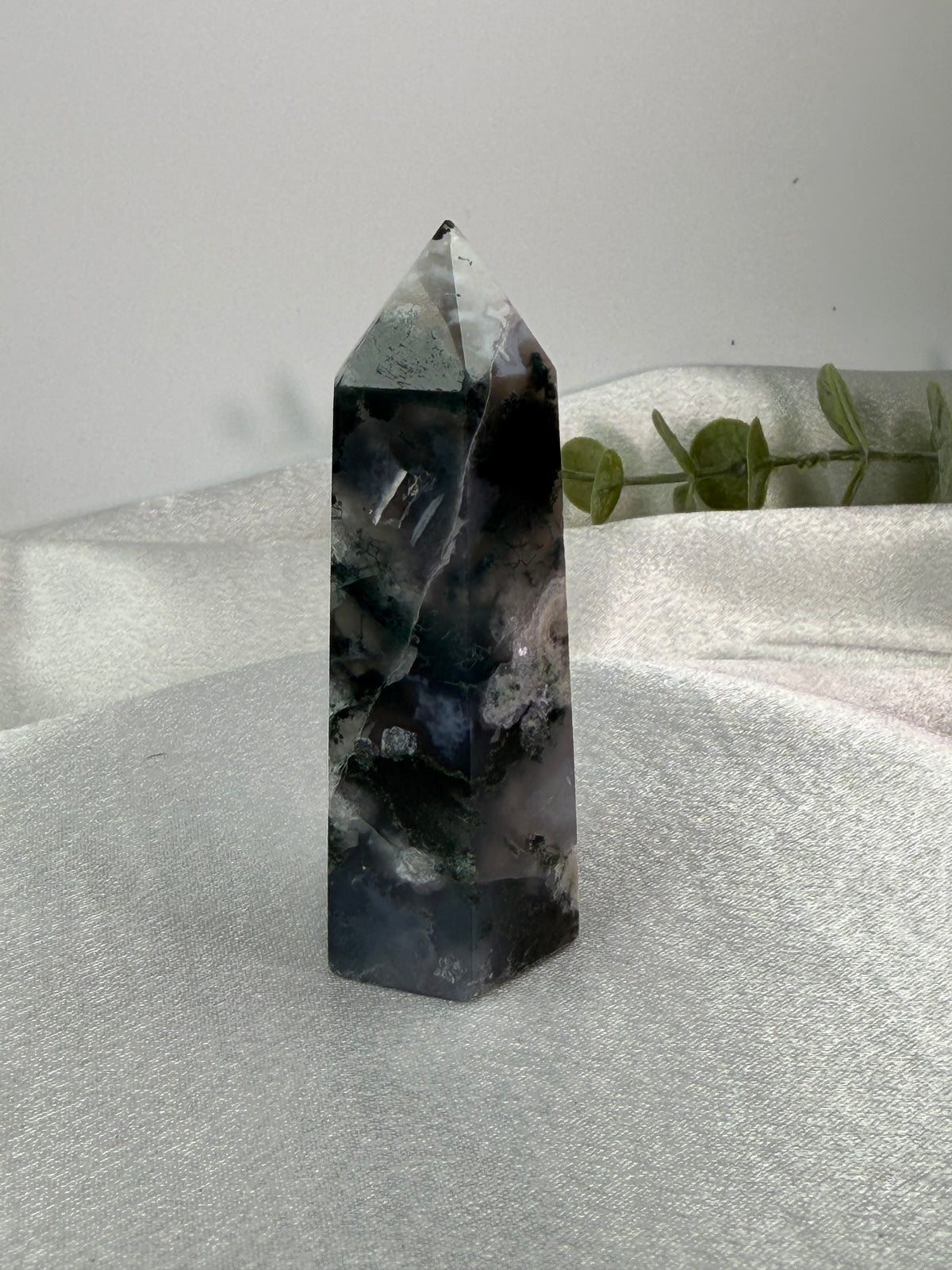 A natural pointed crystal tower, likely amethyst, standing upright on a draped white fabric surface with a soft gray background and a small green plant in the distance.