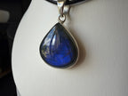 A teardrop-shaped labradorite pendant with a silver bezel setting, displayed on a white jewelry bust. The labradorite exhibits a vibrant blue color with natural iridescent flashes, and the pendant is suspended from a black cord.