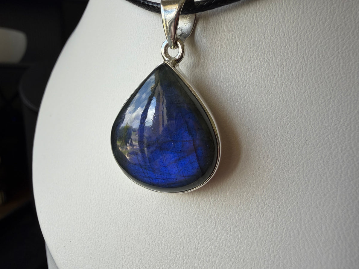 A teardrop-shaped labradorite pendant with a silver bezel setting, displayed on a white jewelry bust. The labradorite exhibits a vibrant blue color with natural iridescent flashes, and the pendant is suspended from a black cord.