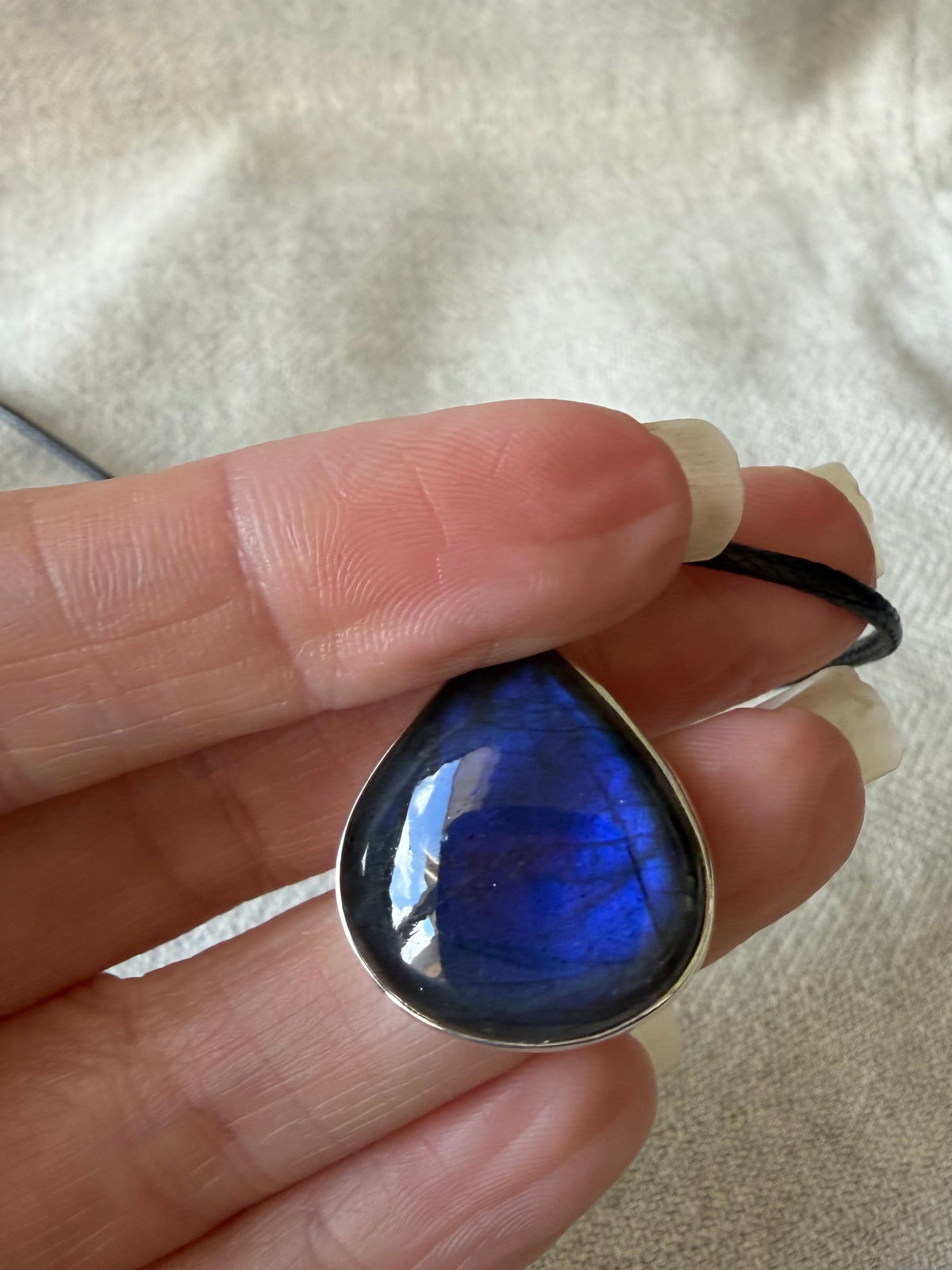 A close-up of a hand holding a teardrop-shaped labradorite pendant necklace. The pendant features a deep blue, iridescent labradorite stone set in a silver bezel, suspended from a black cord.