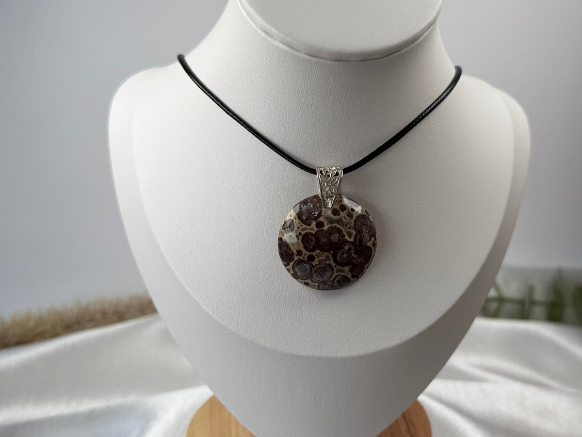 A round poppy jasper pendant necklace with a black cord, displayed on a white jewelry bust stand with a wooden base.