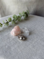 A silver-toned ring with a floral or clover-like raised pattern, displayed on a textured light gray fabric surface next to a pink crystal, a clear quartz crystal, and a green eucalyptus sprig.