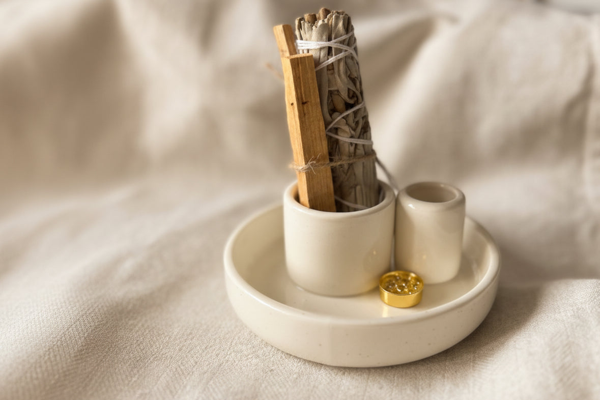 A white ceramic incense holder with a smudge kit bundle and a small gold pendant, set on a draped white linen cloth with a softly blurred background.