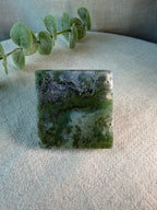 A square polished stone or crystal with marbled green, gray, and white patterns, displayed on a textured beige fabric surface with a green eucalyptus sprig in the background.