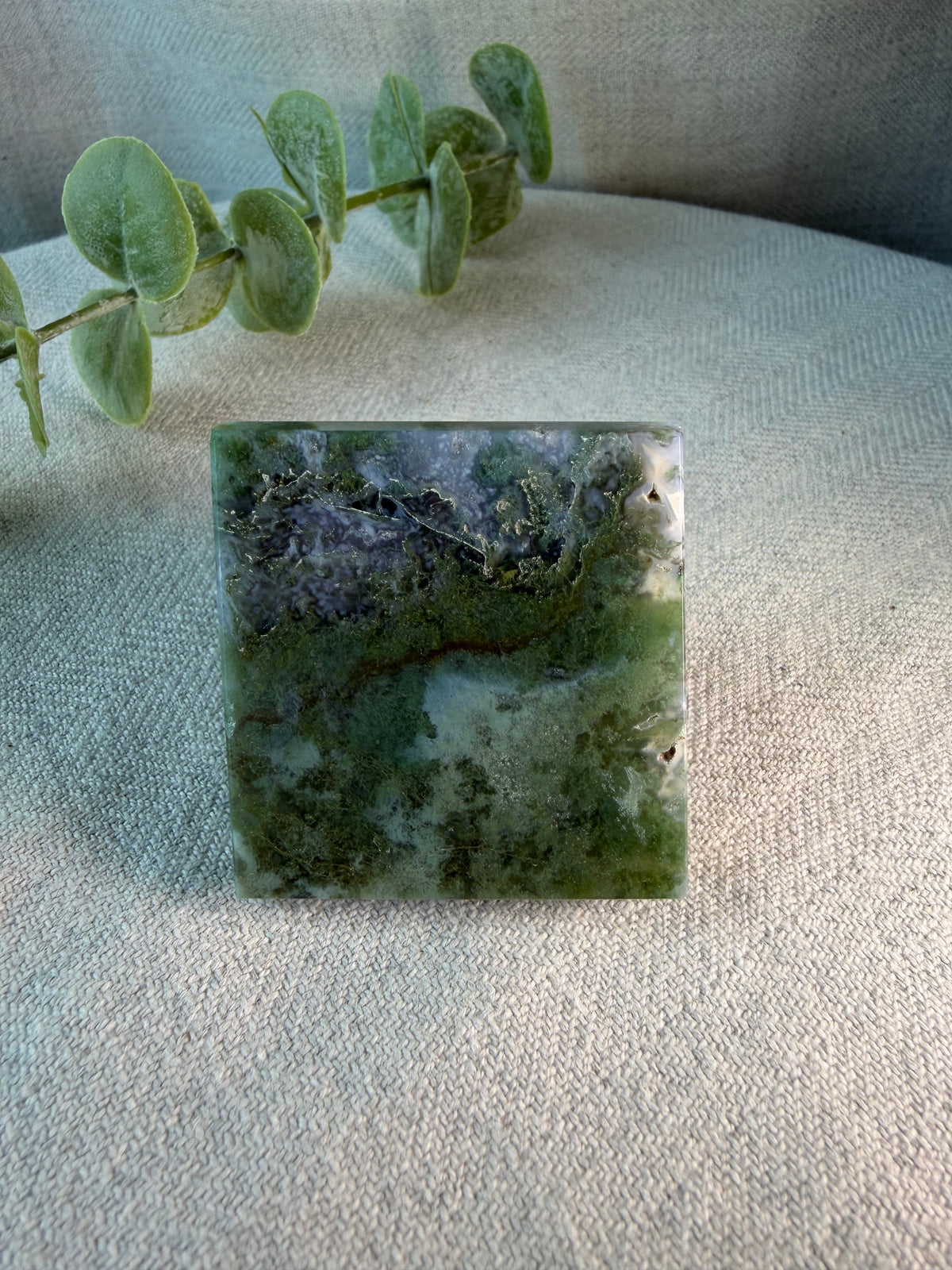 A square polished stone or crystal with marbled green, gray, and white patterns, displayed on a textured beige fabric surface with a green eucalyptus sprig in the background.