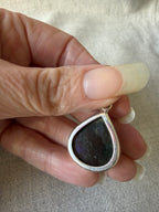 A close-up of a hand holding a teardrop-shaped pendant with a silver-toned border and a dark, iridescent center stone.