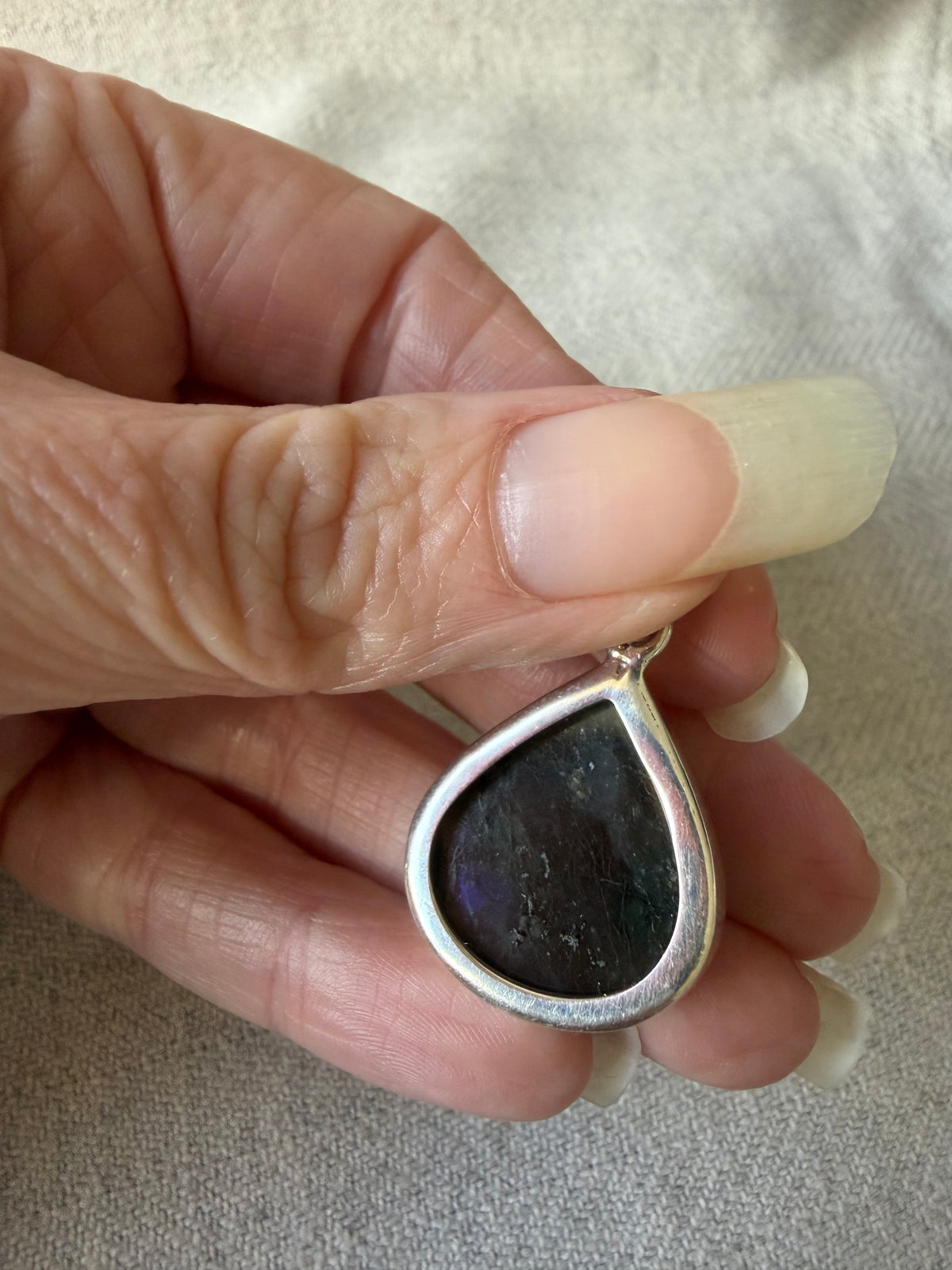 A close-up of a hand holding a teardrop-shaped pendant with a silver-toned border and a dark, iridescent center stone.