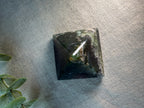 A polished black pyramid crystal tower, likely obsidian, resting on a textured gray fabric surface with green leaves visible in the bottom-left corner.