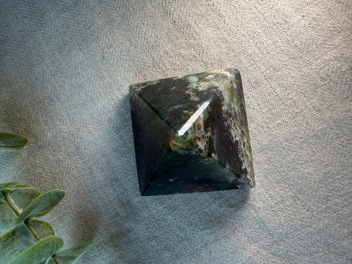A polished black pyramid crystal tower, likely obsidian, resting on a textured gray fabric surface with green leaves visible in the bottom-left corner.
