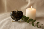 A polished heart-shaped black stone with natural marbled patterns, displayed on a small gold metal stand against a textured off-white fabric background.