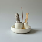 smudge kit A ceramic incense burner set with a shallow round tray, two small cylindrical holders, and a central holder with a gold-toned base, styled with a white sage bundle, a wooden stick, and an incense stick on a textured light gray fabric backdrop.