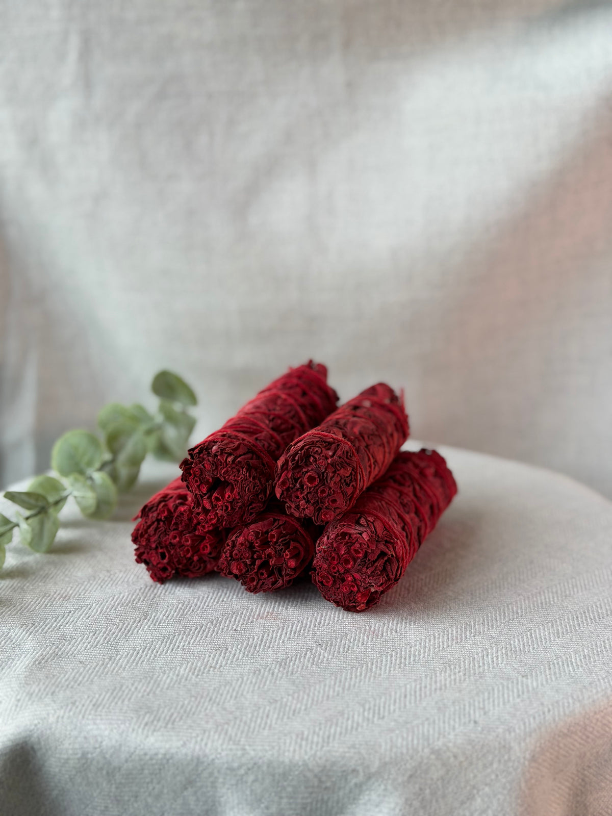 Four dried red sage smudge sticks arranged on a light beige fabric surface, with a sprig of green eucalyptus leaves on the left and a soft neutral fabric backdrop behind.