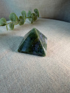A polished green crystal pyramid stone resting on a textured beige fabric surface, with a green eucalyptus branch in the background.