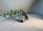 A polished crystal pyramid with natural green and black banded stone patterns, resting on a textured beige fabric surface next to a green eucalyptus sprig.