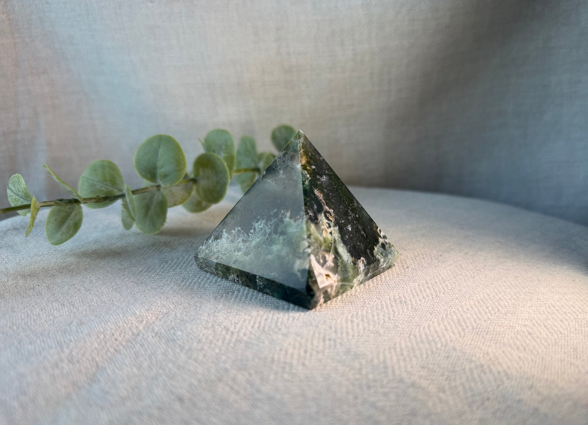 A polished crystal pyramid with natural green and black banded stone patterns, resting on a textured beige fabric surface next to a green eucalyptus sprig.
