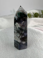 A polished natural crystal tower with a pointed apex, featuring marbled patterns of black, gray, white, and green, displayed on a textured light gray fabric surface with a soft-focus background.