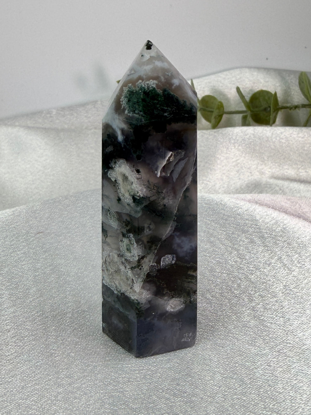 A polished natural crystal tower with a pointed apex, featuring marbled patterns of black, gray, white, and green, displayed on a textured light gray fabric surface with a soft-focus background.