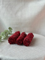 Three rolled bundles of deep red dried flower petals, likely rose petals, arranged on a light beige textured fabric surface next to a green eucalyptus sprig.
