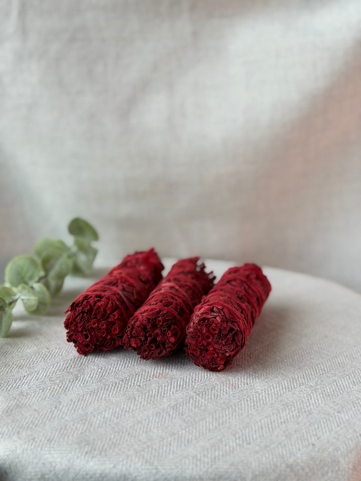 Three rolled bundles of deep red dried flower petals, likely rose petals, arranged on a light beige textured fabric surface next to a green eucalyptus sprig.