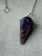 Faceted purple amethyst crystal pendant necklace on a silver chain, displayed on a textured gray fabric background.