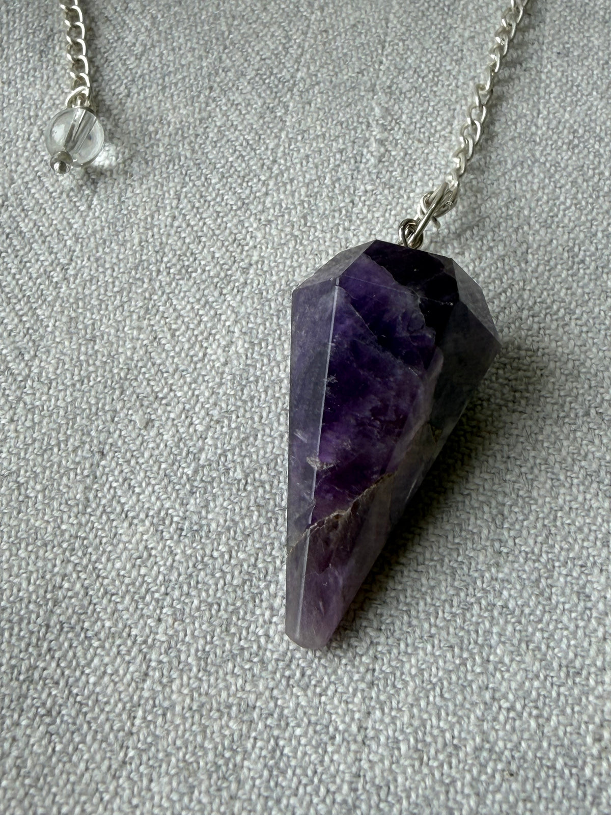Faceted purple amethyst crystal pendant necklace on a silver chain, displayed on a textured gray fabric background.