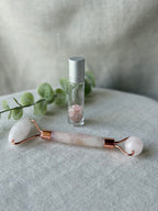 A rose quartz facial roller with rose gold metal accents lies on a textured light gray fabric surface next to a small glass roller bottle with a silver cap and a sprig of green eucalyptus.
