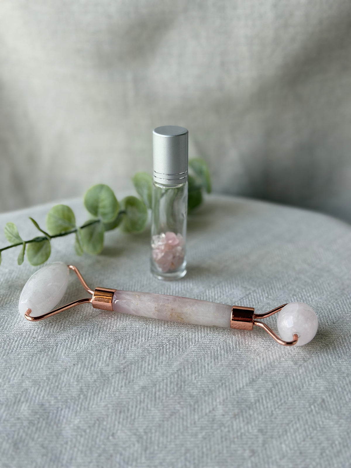 A rose quartz facial roller with rose gold metal accents lies on a textured light gray fabric surface next to a small glass roller bottle with a silver cap and a sprig of green eucalyptus.