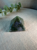 A polished green crystal pyramid with natural inclusions, displayed on a textured beige fabric surface with a green eucalyptus branch in the background.