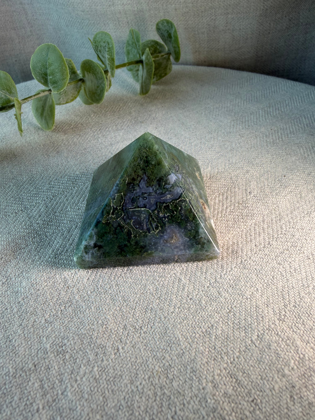 A polished green crystal pyramid with natural inclusions, displayed on a textured beige fabric surface with a green eucalyptus branch in the background.