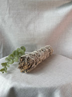 A dried sage smudge bundle wrapped in white string, resting on a draped light beige fabric surface next to a small green eucalyptus sprig.