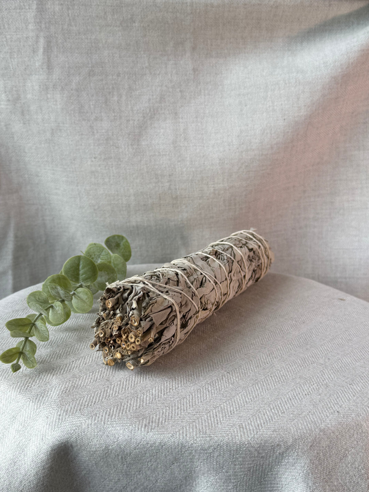 A dried sage smudge bundle wrapped in white string, resting on a draped light beige fabric surface next to a small green eucalyptus sprig.