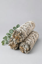 Two dried sage smudge bundles resting on a light beige textured fabric surface, with a small green sprig nearby and a soft window light casting gentle shadows.