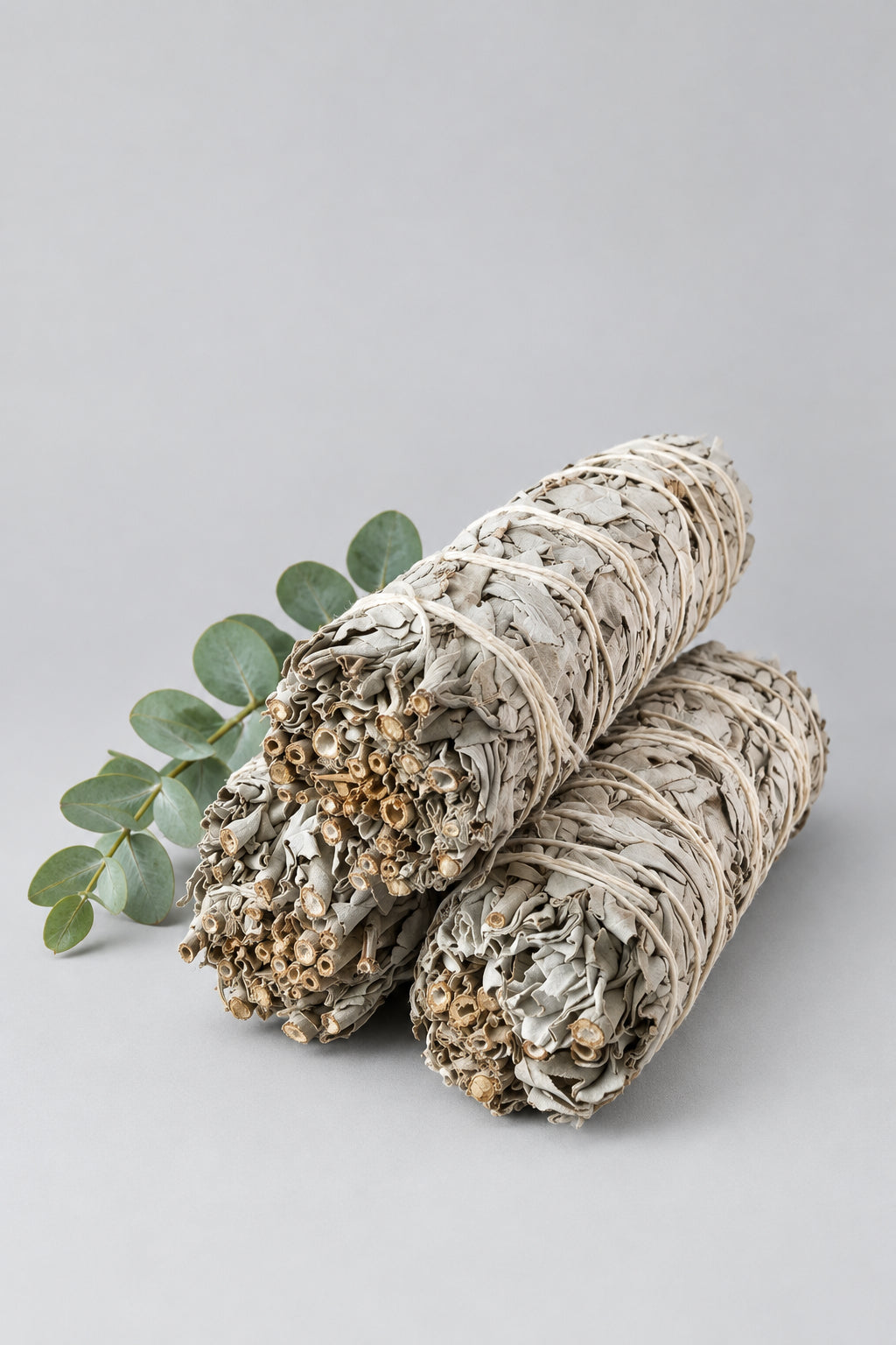 Two dried sage smudge bundles resting on a light beige textured fabric surface, with a small green sprig nearby and a soft window light casting gentle shadows.
