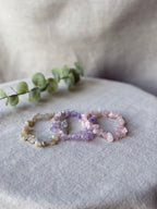 Three beaded stretch bracelets made of natural gemstone chips in beige, purple, and pink, arranged on a textured light gray fabric surface with a green eucalyptus sprig in the background.