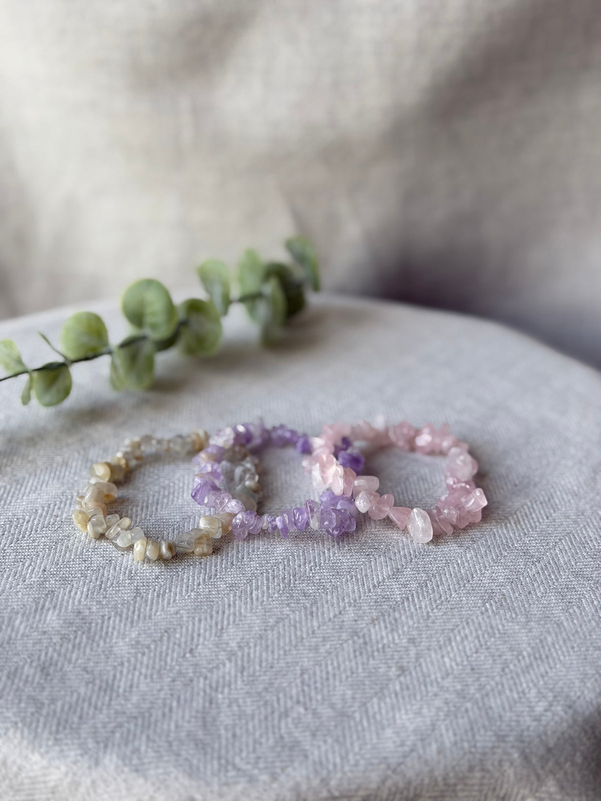 Three beaded stretch bracelets made of natural gemstone chips in beige, purple, and pink, arranged on a textured light gray fabric surface with a green eucalyptus sprig in the background.