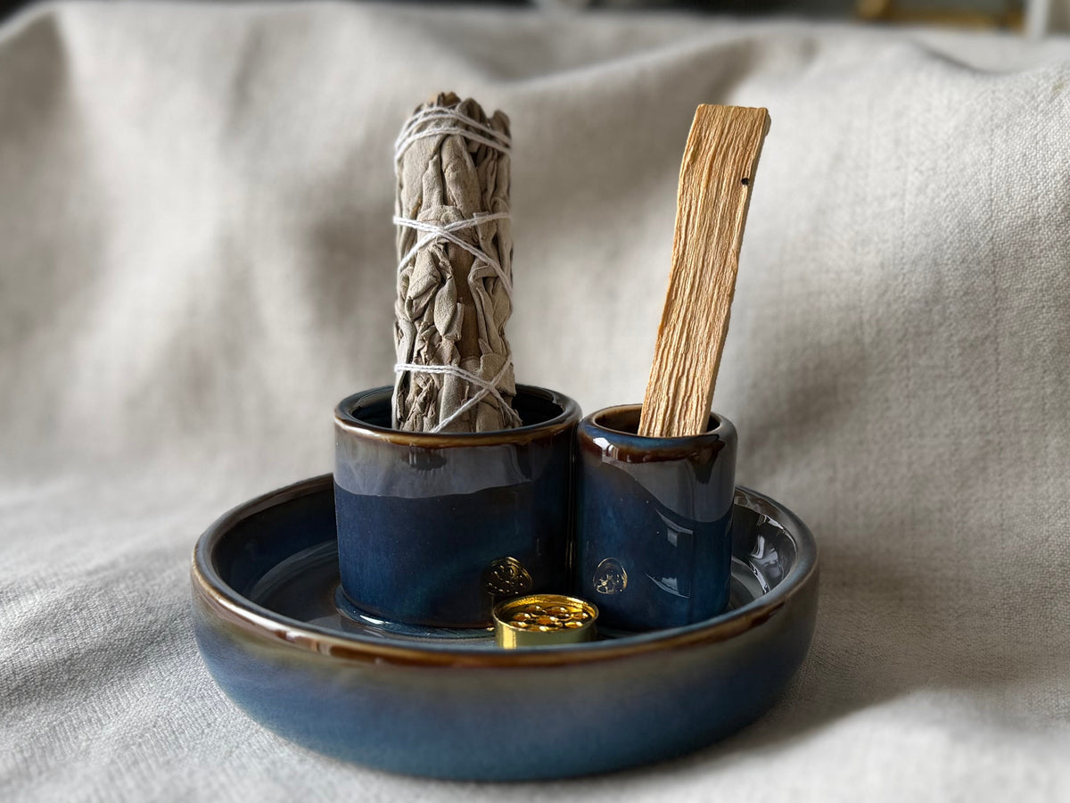Smudge Kit A small ceramic set with a round shallow dish and two small cups in a deep blue glaze with a gold-brown rim, holding a sage bundle and a wooden stick, placed on a draped white linen cloth.
