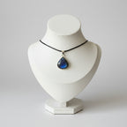A silver-toned teardrop pendant necklace with a deep blue and black starry sky design, displayed on a cream-colored jewelry bust on a wooden stand.