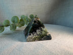 A triangular crystal moss agate pyramid stone with natural green and black marbled patterns, styled on a textured beige fabric surface next to a green eucalyptus sprig.